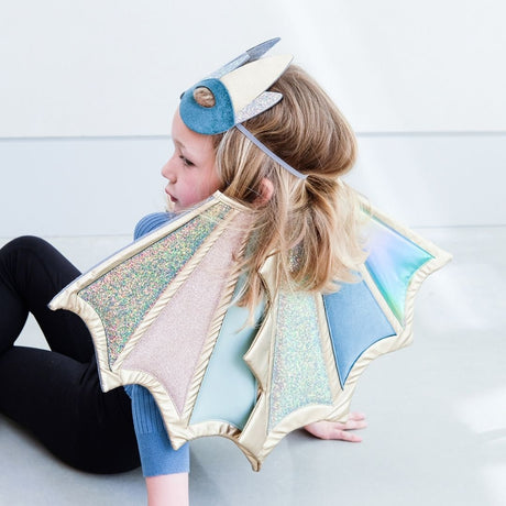 Pige sidder, set bagfra, med dragemaske i panden og vinger på ryggen i pastelfarver med glitter og guld fra Mimi & Lula fås hos Nordic Simply