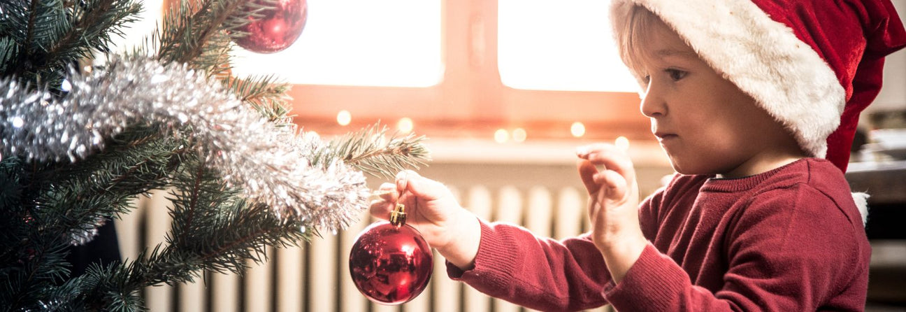 barn 3-5 år med rød nissehue og rød bluse hænger rød julekugle på pyntet juletræ, gør julen ekstra magisk for børn og børnebørn med adventsgaver fra Nordic Simply