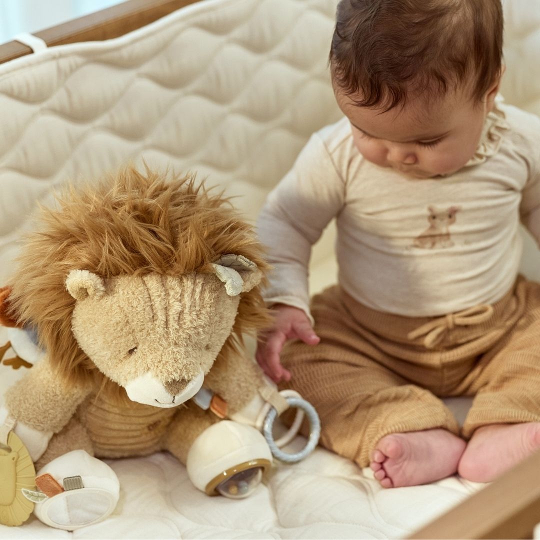 Baby sidder på lyst tæppe og ser på løve aktivitetsbamse. En sød bamse med masser at udforske fra nyfødt og op. Little Dutch. 
