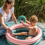 mor leger med dreng, der sidder i lille rundt badebassin med to ringe, en grøn og en orange med hvide striber, bassinet har en diameter på 80 centimeter.