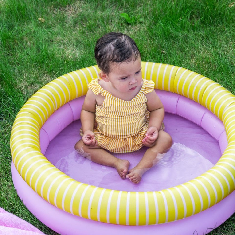 Pige sidder i lille rundt badebassin med to ringe, en rosa og en gul med hvide striber, bassinet har en diameter på 80 centimeter og er derfor til små børn under opsyn.