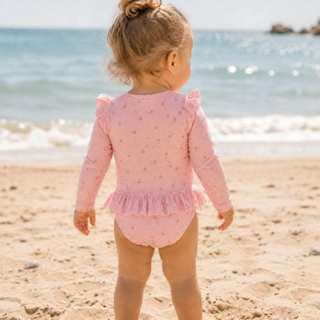 pige på strand i rosa badedragt med lange ærmer og pink motiver af blomster, badedragt med UV40 beskyttelse, set bagfra.