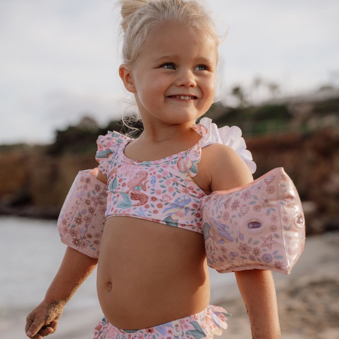 Pige på strand med rosa armvinger, med havfruemotiver, på.
