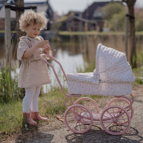 Pige ude med barnevogn til dukke. Med rosa stel og hvidt betræk med små blomster. Flot dukkevogn til ture med dukke både ude og inde, fra Little Dutch, fås hos Nordic Simply