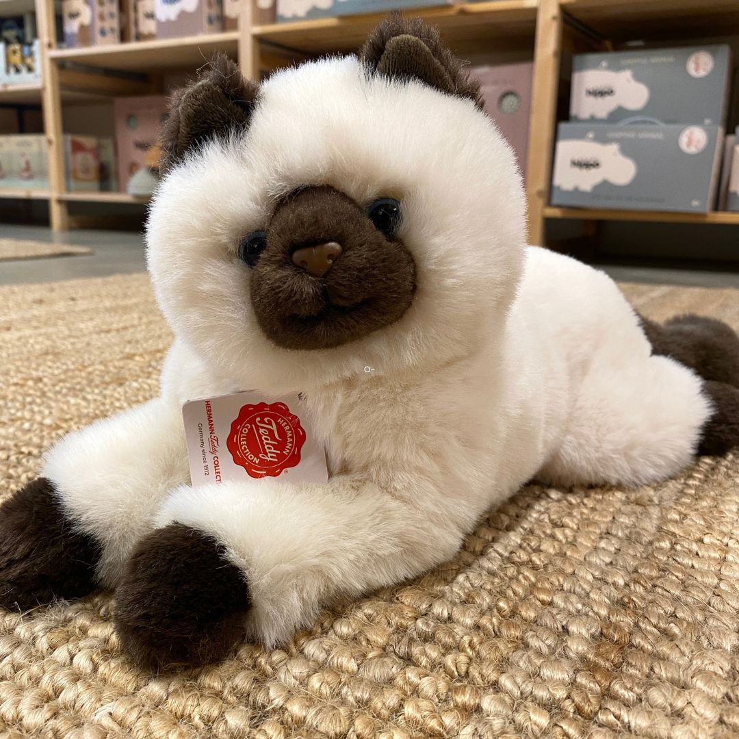 Hvid og sort Birma kattebamse med store blå øjne, og busket hale, ligger på lyst gulvtæppe, hos Nordic Simply