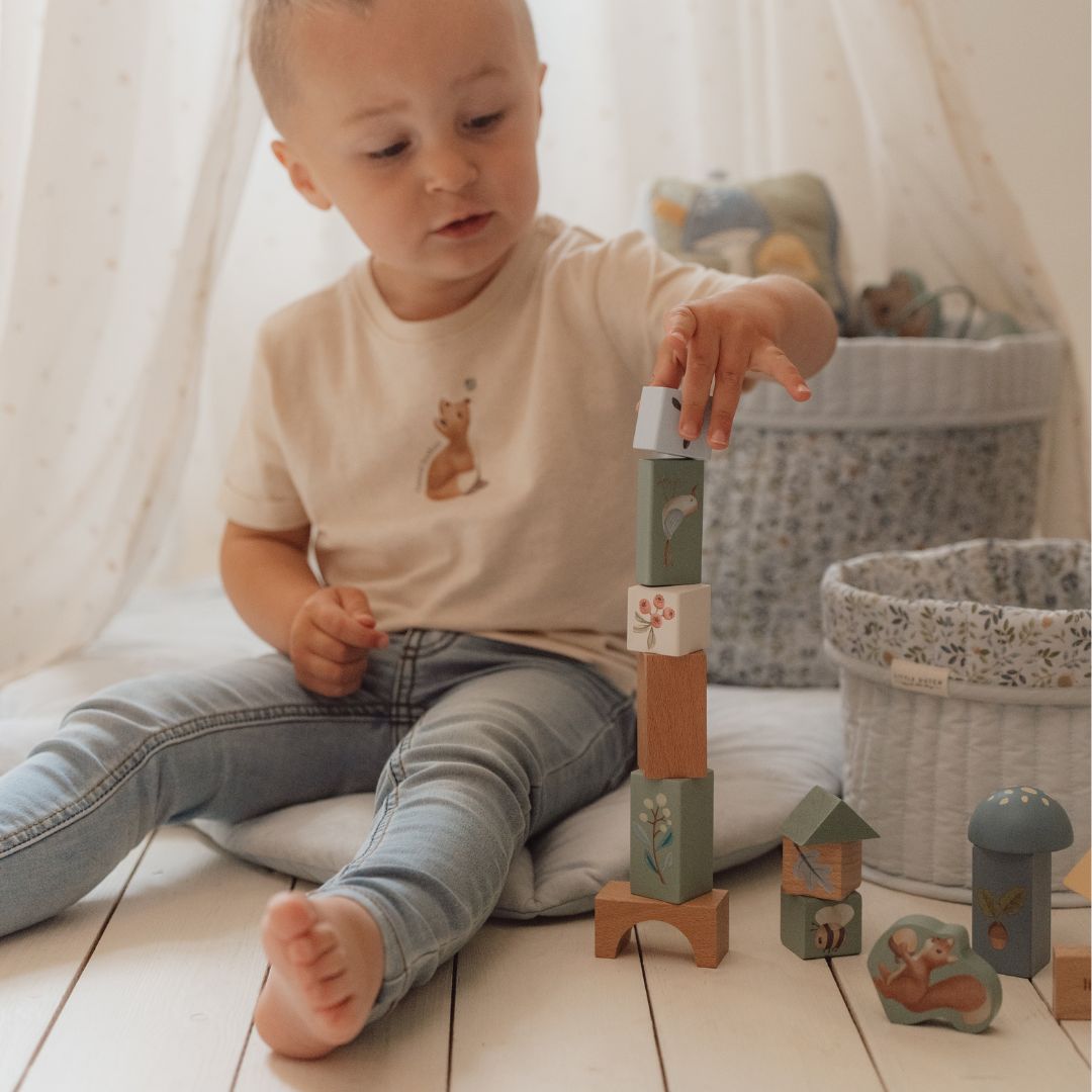 Lille dreng bygger med farverige træklodser, iført en T-shirt med dyremotiv, i en hyggelig indendørs legestue.