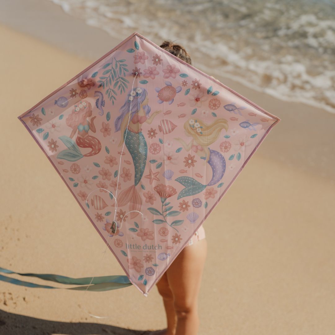 barn på strand holder rosa drage med motiver af havfruer op foran ansigtet.