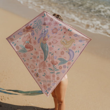 barn på strand holder rosa drage med motiver af havfruer op foran ansigtet.