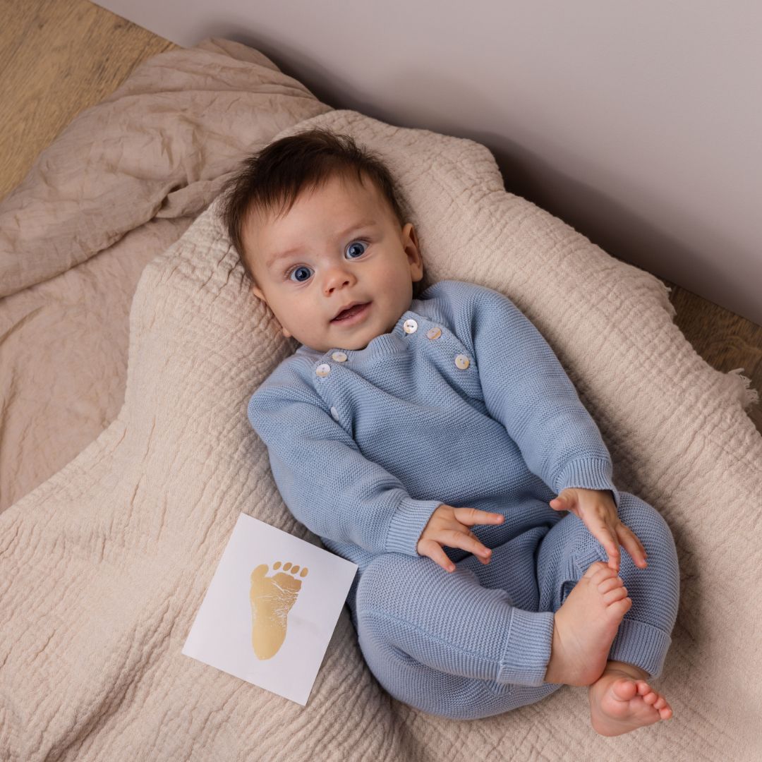 fodaftryk i guld ved baby, der ligger på lyst tæppe i blå dragt, med bare fødder, set hos Nordic Simply