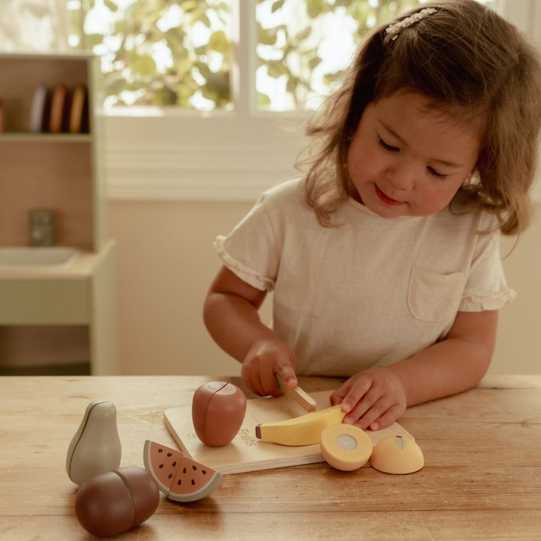 Pige skærer banan med kniv i træ på skærebræt i træ fra frugtsæt med seks frugter til dit barn, fra Little Dutch.