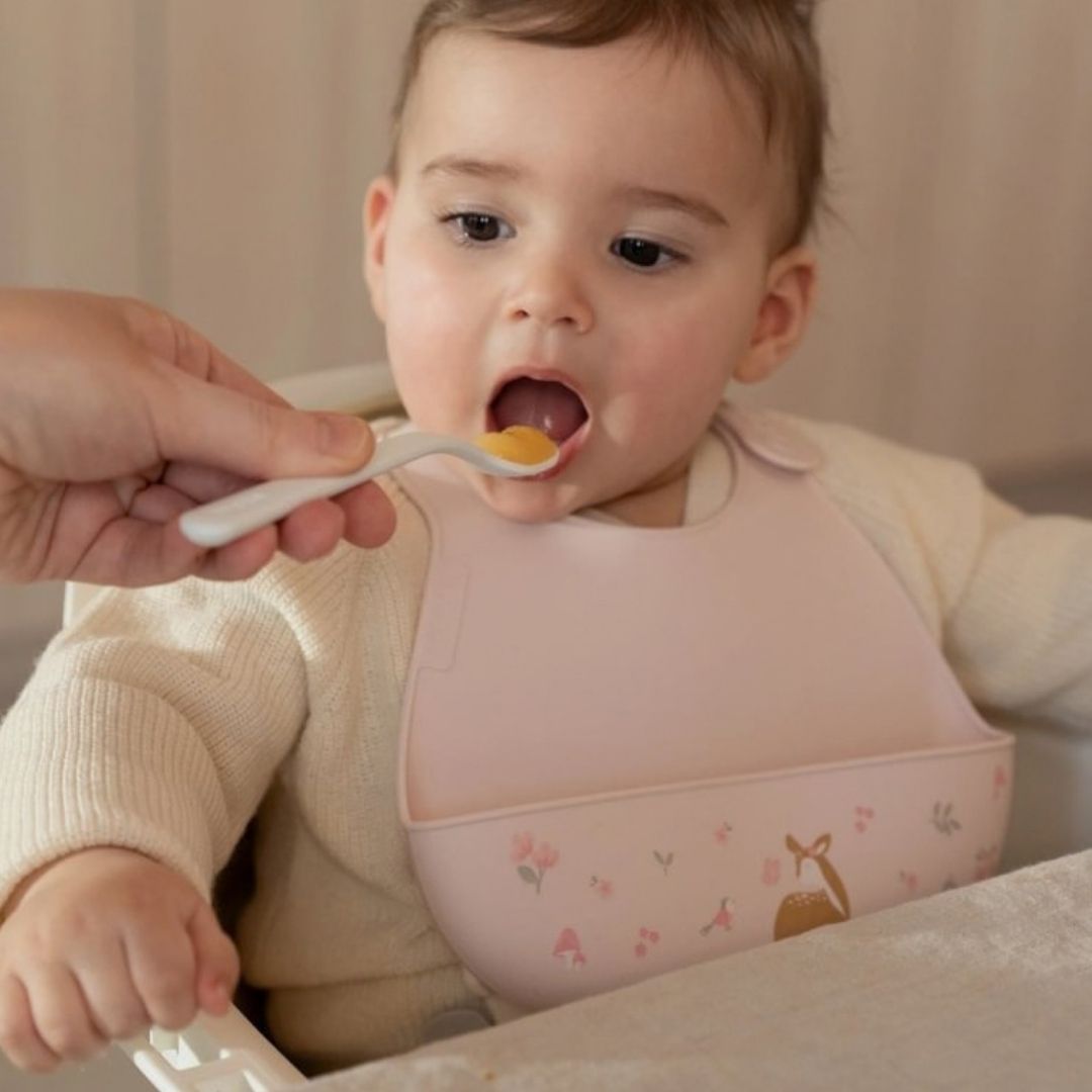 baby, med rosa hagesmæk med opsamlingslomme og sødt motiv af rådyr, får mad med ske.