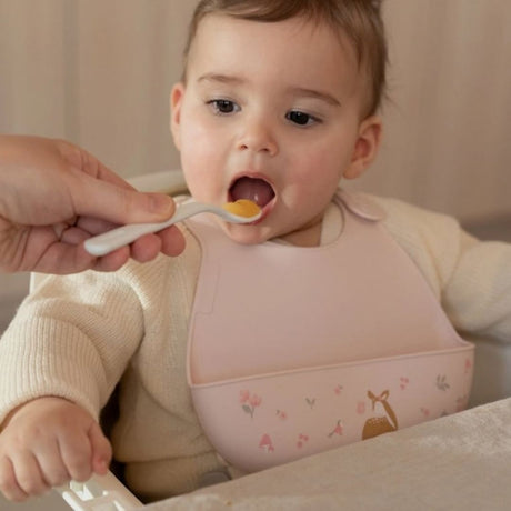 baby, med rosa hagesmæk med opsamlingslomme og sødt motiv af rådyr, får mad med ske.