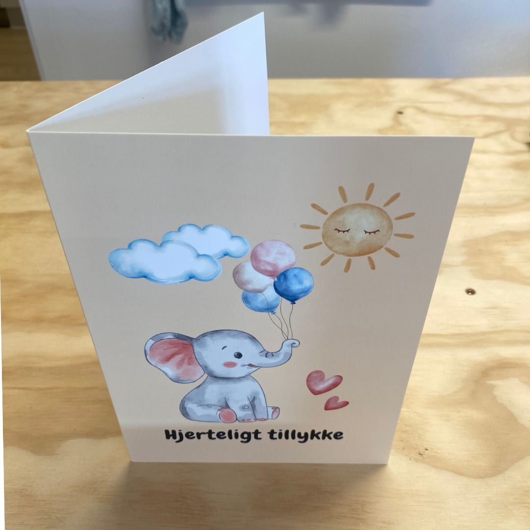 lykønskningskort A5 med motiver af elefant med balloner og teksten hjerteligt tillykke. Flot dobbeltkort til dåb, fødselsdag og andre anledninger for børn. Her set åbnet på bord.