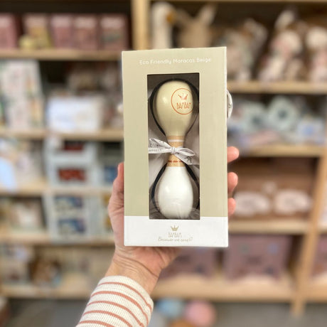 Hånd holder gaveæske med beige og hvid maracas i træ fra BamBam med rundt BamBam logo. Dejligt lille instrument til barn fra 18 måneder, fås hos Nordic Simply.