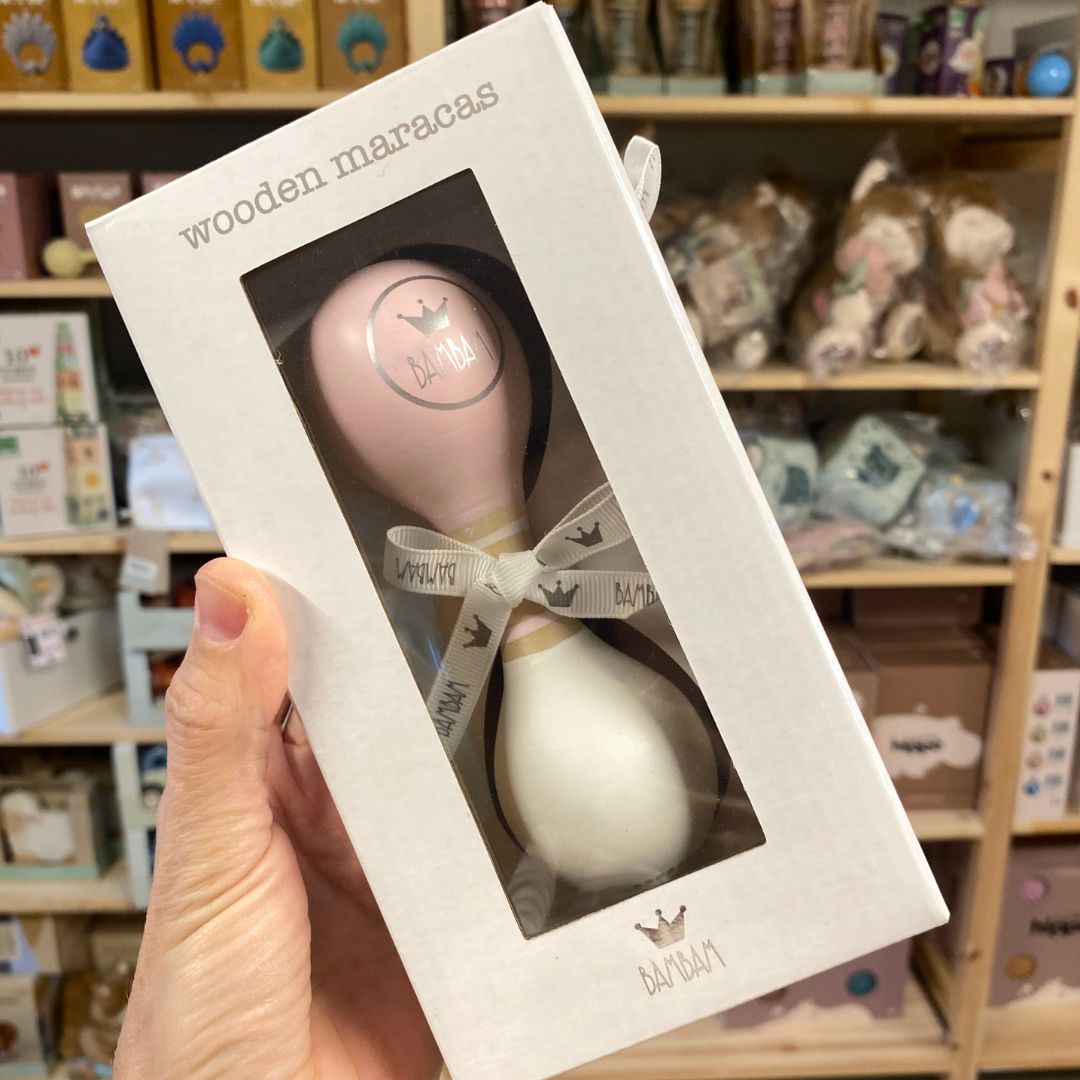 Hånd holder gaveæske medrosa og hvid maracas i træ fra BamBam med rundt BamBam logo. Dejligt lille instrument til barn fra 18 måneder, fås hos Nordic Simply.