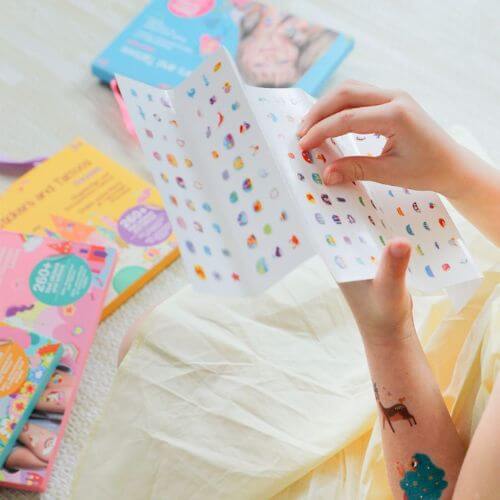 Child exploring colorful nail stickers and tattoos from a floral-themed set, surrounded by fun activity books.