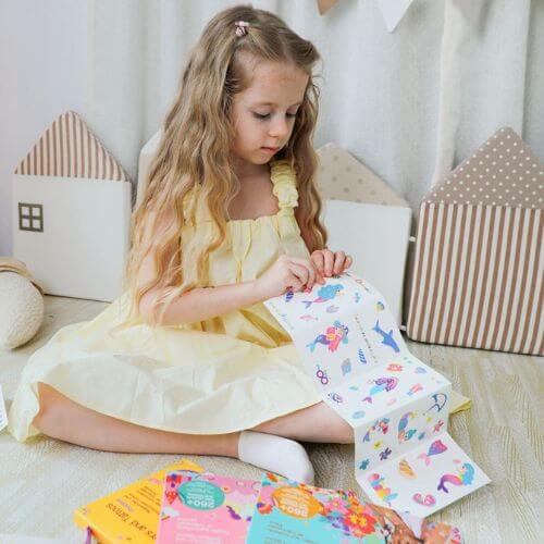 Little girl in a yellow dress happily exploring colorful nail stickers and tattoos on a cozy playroom floor.