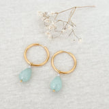 Små guld hoops øeringe med turkis amazonit dråbe, facetslebet, set på lys baggrund med lille hvid blomst ovenover.