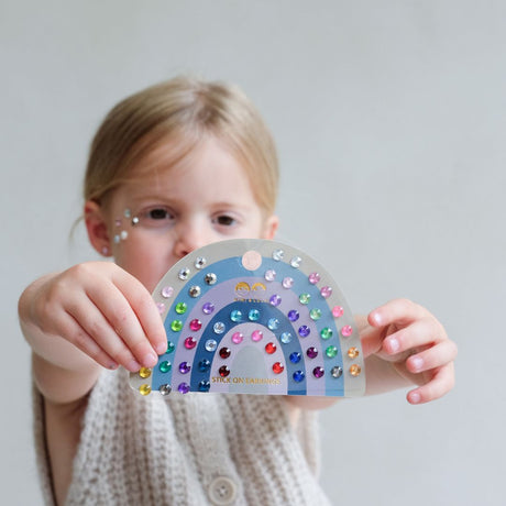 Pige holder ark, udformet som regnbue frem, med 30 par ørestickers i multifarver til børn. Øreringene klistres på, så du behøver ikke huller i ørerne.