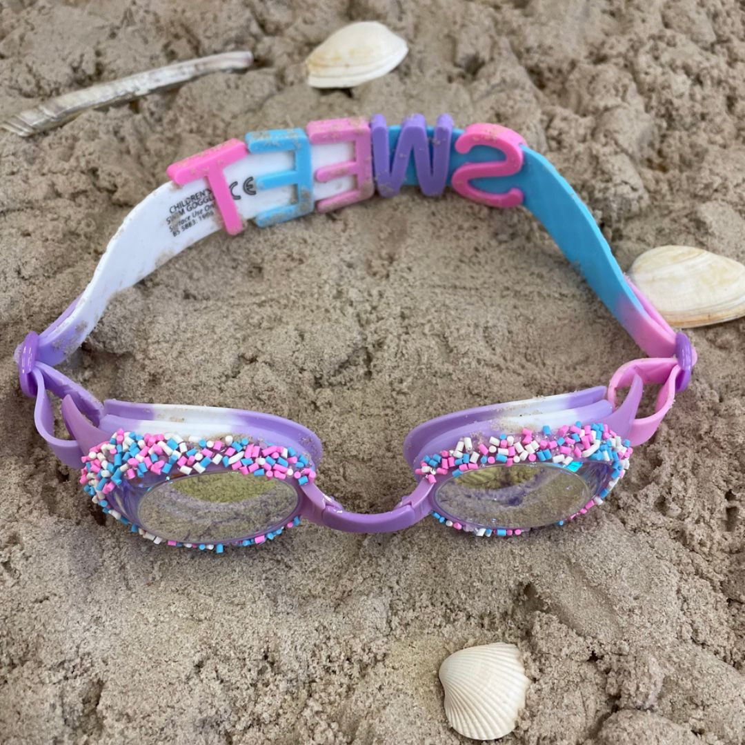 lilla svømmebriller dekoreret med krymmel og konfetti fra Bling20, her set i sand med hvide muslinger omkring