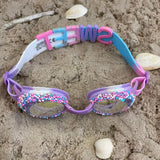 lilla svømmebriller dekoreret med krymmel og konfetti fra Bling20, her set i sand med hvide muslinger omkring